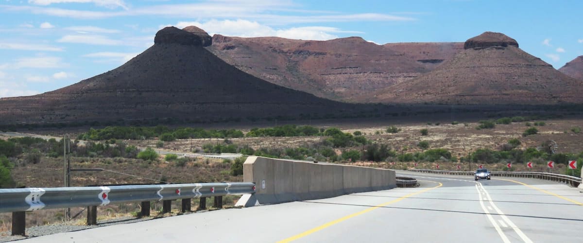 A highway with guardrails, a mountain range, and sparse vegetation.