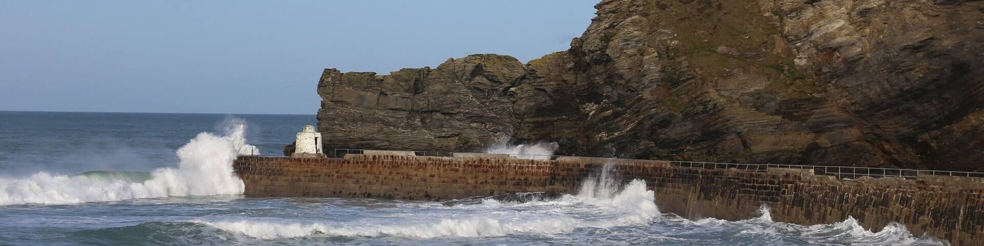 A rocky coastline with waves crashing against a sea wall and a lighthouse on top of a cliff.