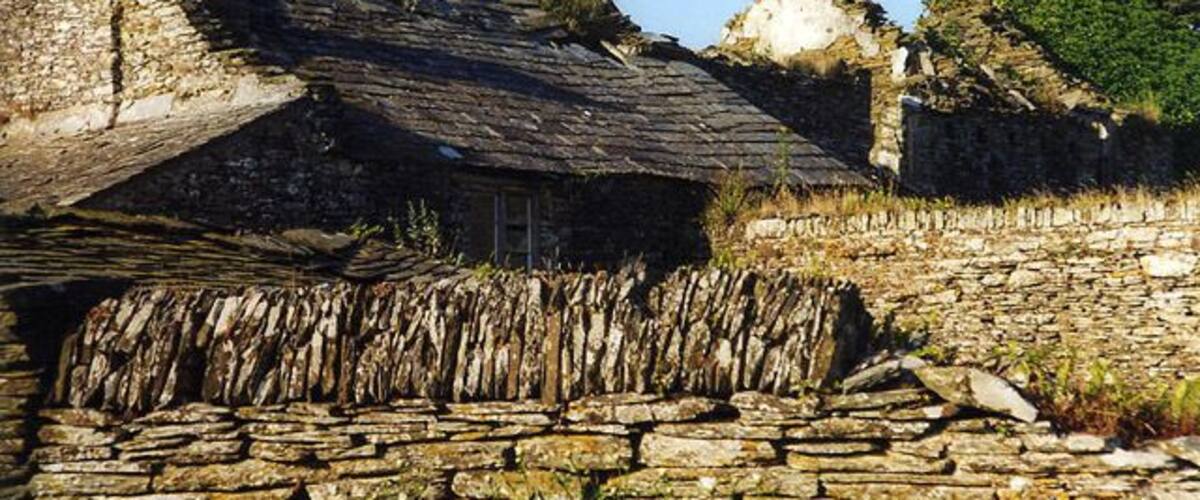 A stone structure with a sloped roof, surrounded by stone walls and vegetation.