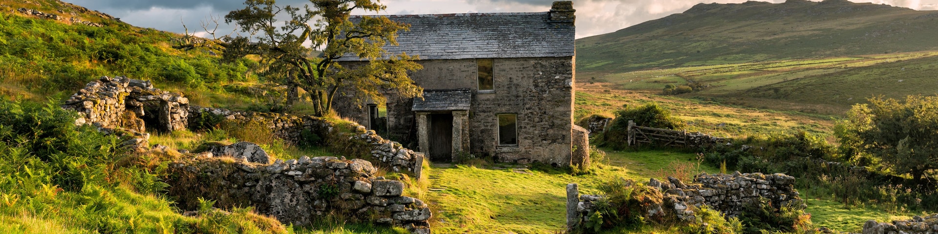 Une maison en pierre, coiffée d’un toit de chaume, entourée de collines verdoyantes et sous un ciel nuageux.