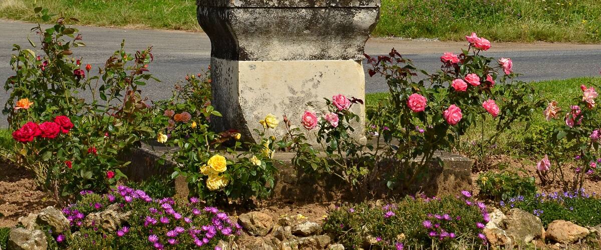 A weathered stone cross in a rural setting with a road, flowering plants, and a field in the background.