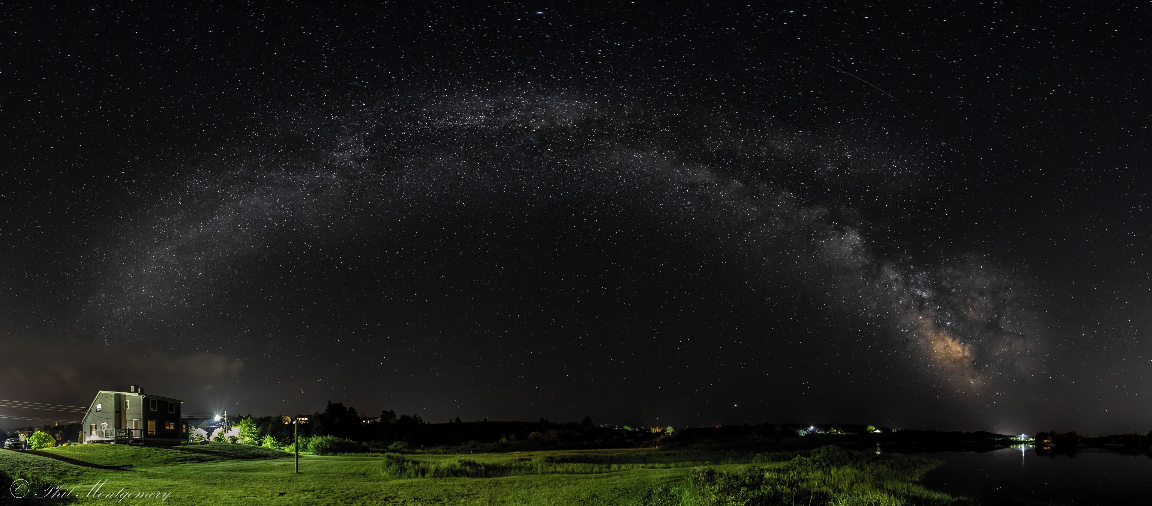 A starry night sky over a grassy field with a house and a lake.