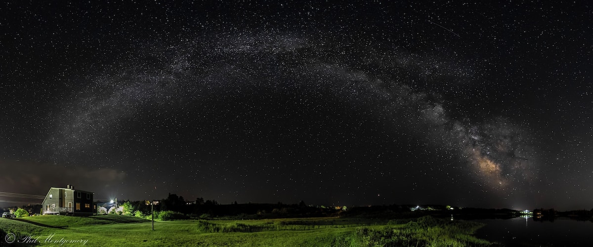 A starry night sky over a grassy field with a house and a lake.