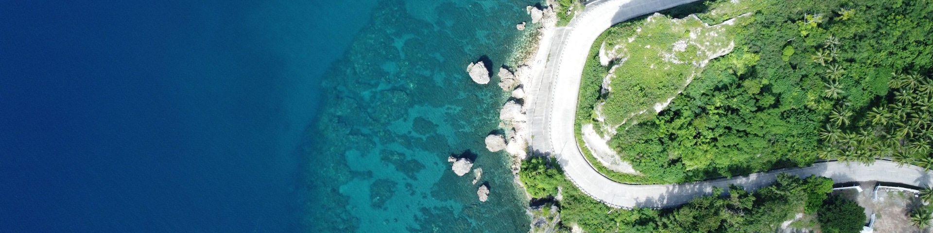 Uma estrada costeira serpenteia por uma paisagem verdejante, com águas azul-claras e afloramentos rochosos.