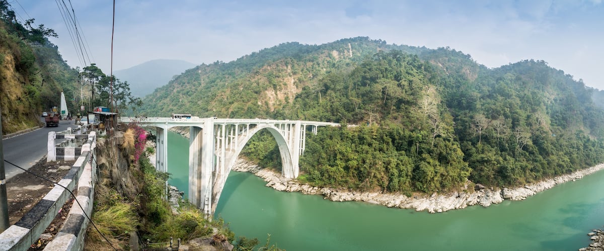 A white bridge over a green river, surrounded by forested hills.