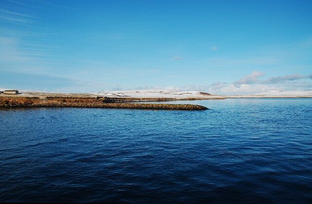 A calm body of water, a rocky shoreline, and a distant snow-covered landscape under a clear sky.