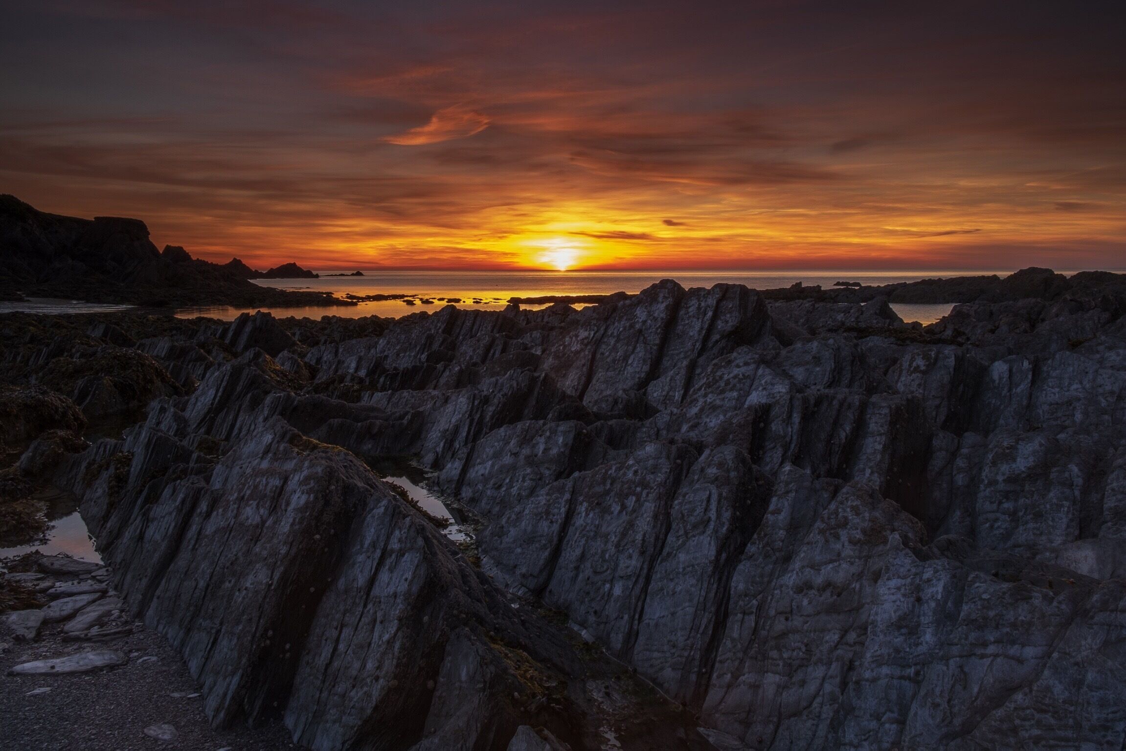 A rocky coastline at sunset with a visible sun near the horizon.