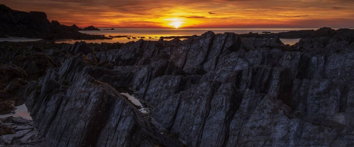 A rocky coastline at sunset with a visible sun near the horizon.
