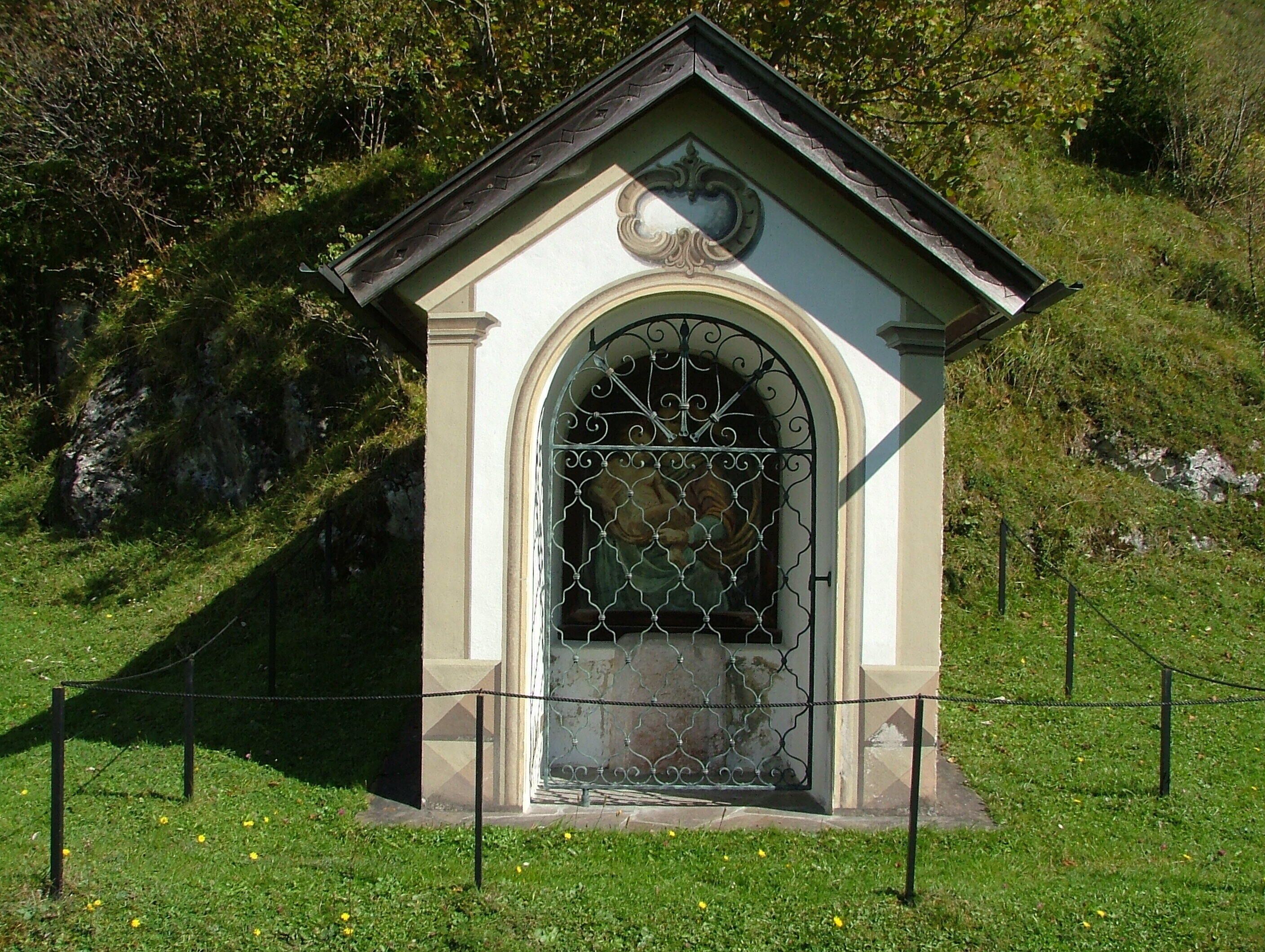 A small, ornate building with a decorative archway and a metal gate, surrounded by a chain-link fence and grass.