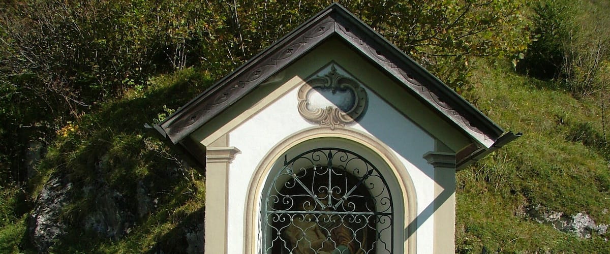 A small, ornate building with a decorative archway and a metal gate, surrounded by a chain-link fence and grass.