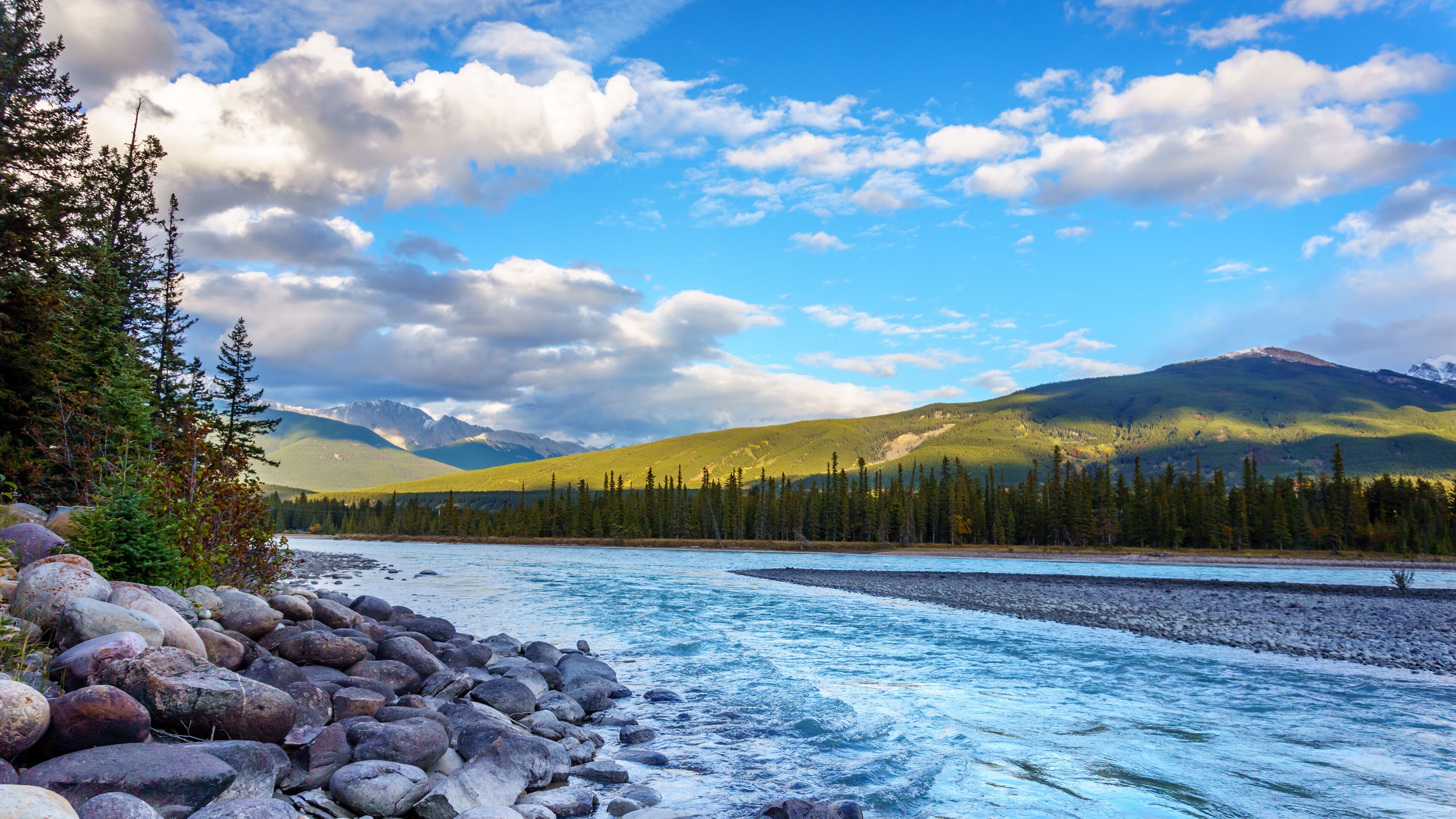 A river with a rocky shoreline, surrounded by dense forests and mountains under a partly cloudy sky.