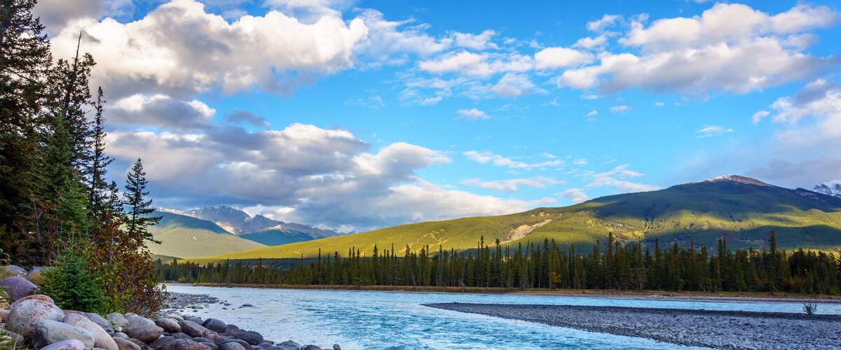 A river with a rocky shoreline, surrounded by dense forests and mountains under a partly cloudy sky.