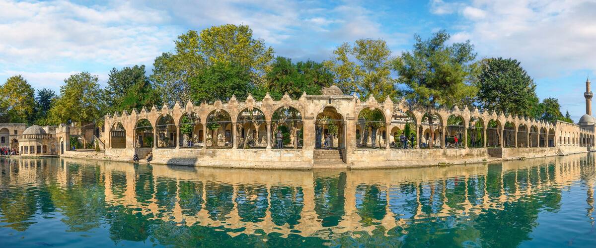A historic stone bridge with arches over a calm river, surrounded by trees and a mosque with a minaret in the background.