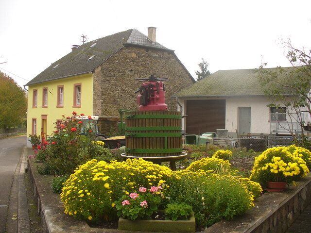 A yellow house with a stone roof, a green and red water tower, and flower beds with yellow and pink flowers.