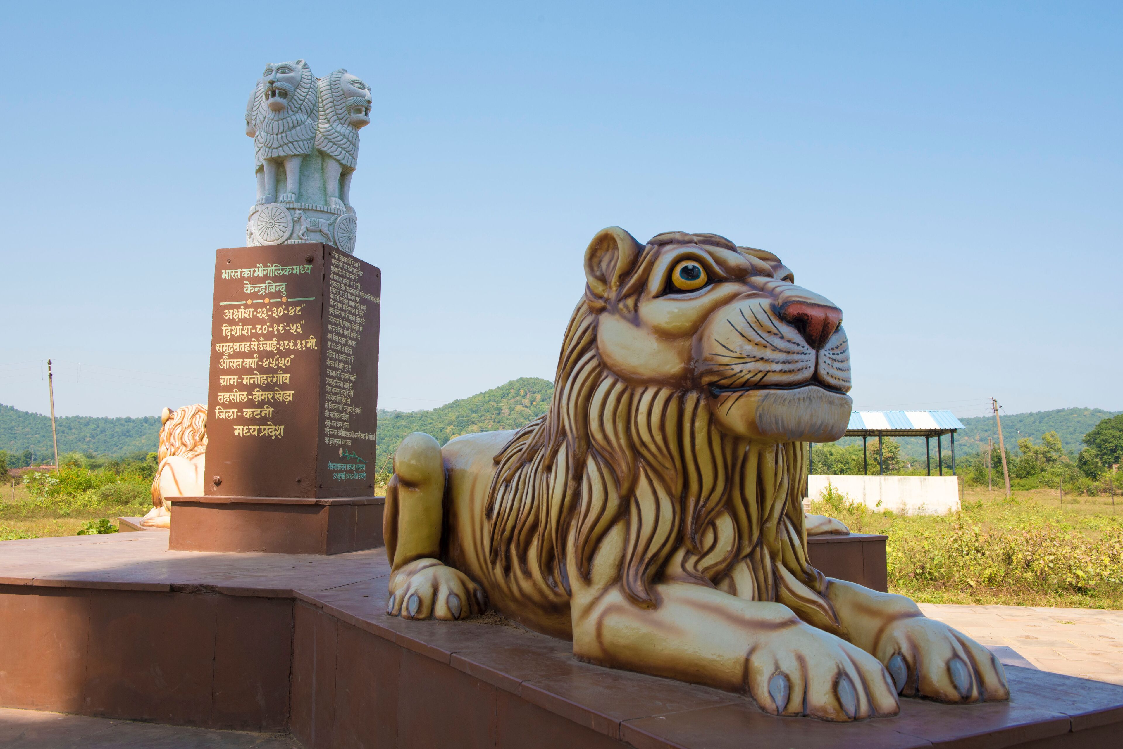 A large bronze statue of a tiger and a smaller stone statue of a lion, both with inscriptions in Hindi, set against a backdrop of green hills and clear sky.