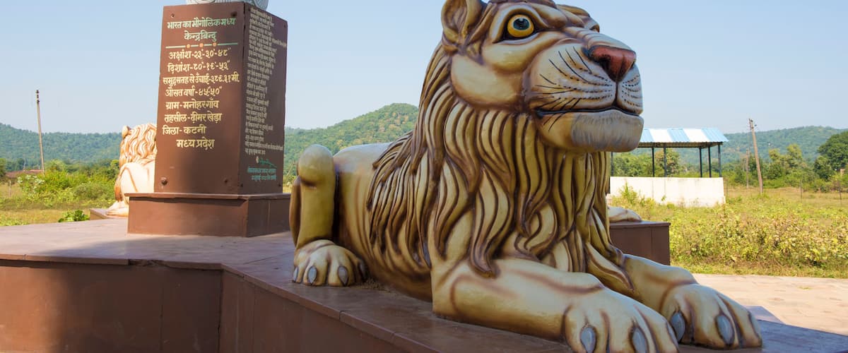 A large bronze statue of a tiger and a smaller stone statue of a lion, both with inscriptions in Hindi, set against a backdrop of green hills and clear sky.