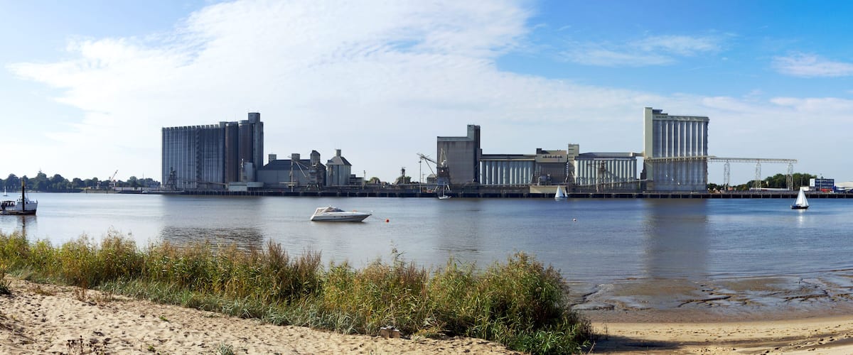 A river with boats, a sandy beach, and industrial buildings in the background.