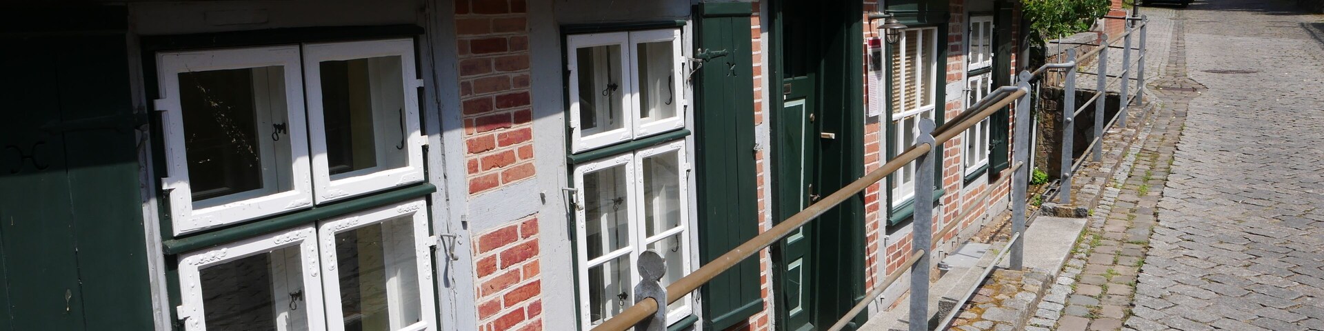 Uma fileira de edifícios tradicionais de tijolo, com telhados de telha vermelha e portejas verdes, uma rua de calçada e um corrimão de metal.