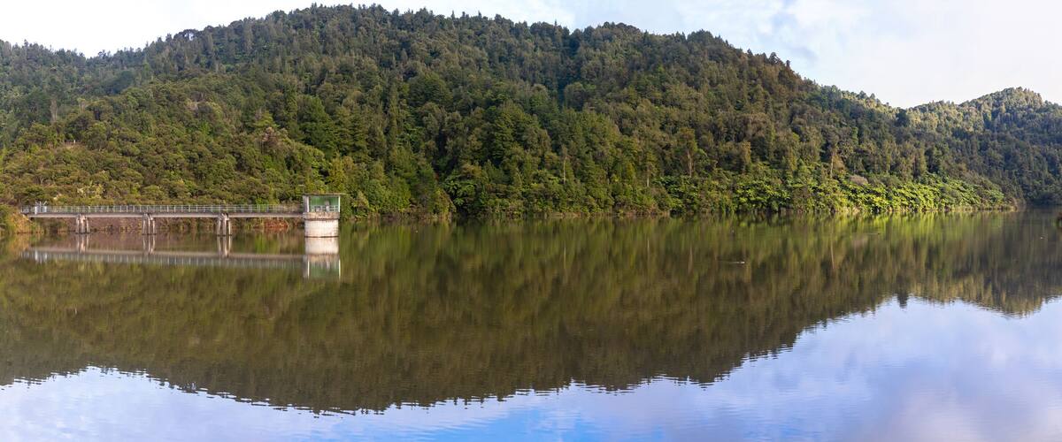 A calm lake with a bridge and dense forested hills in the background.