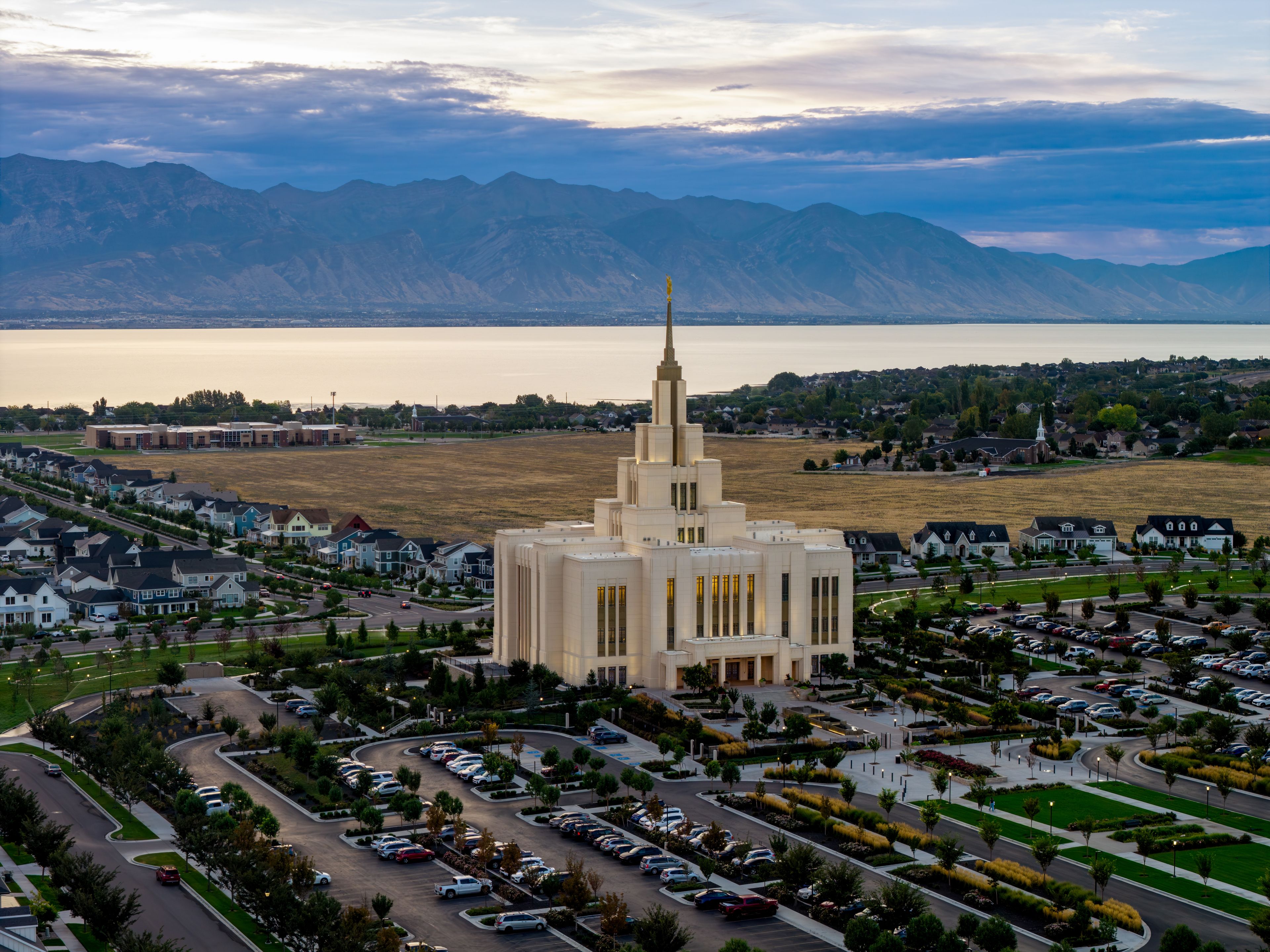 A large building with a tall spire, surrounded by a parking lot and residential houses, with mountains in the background.