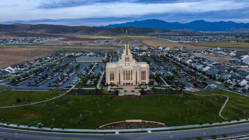 Saratoga Springs, Utah, United States - 22 August 2026: Aerial view of Saratoga Springs Utah Temple glowing during the blue hour, surrounded by lawns, parking lots, and residential neighborhoods.