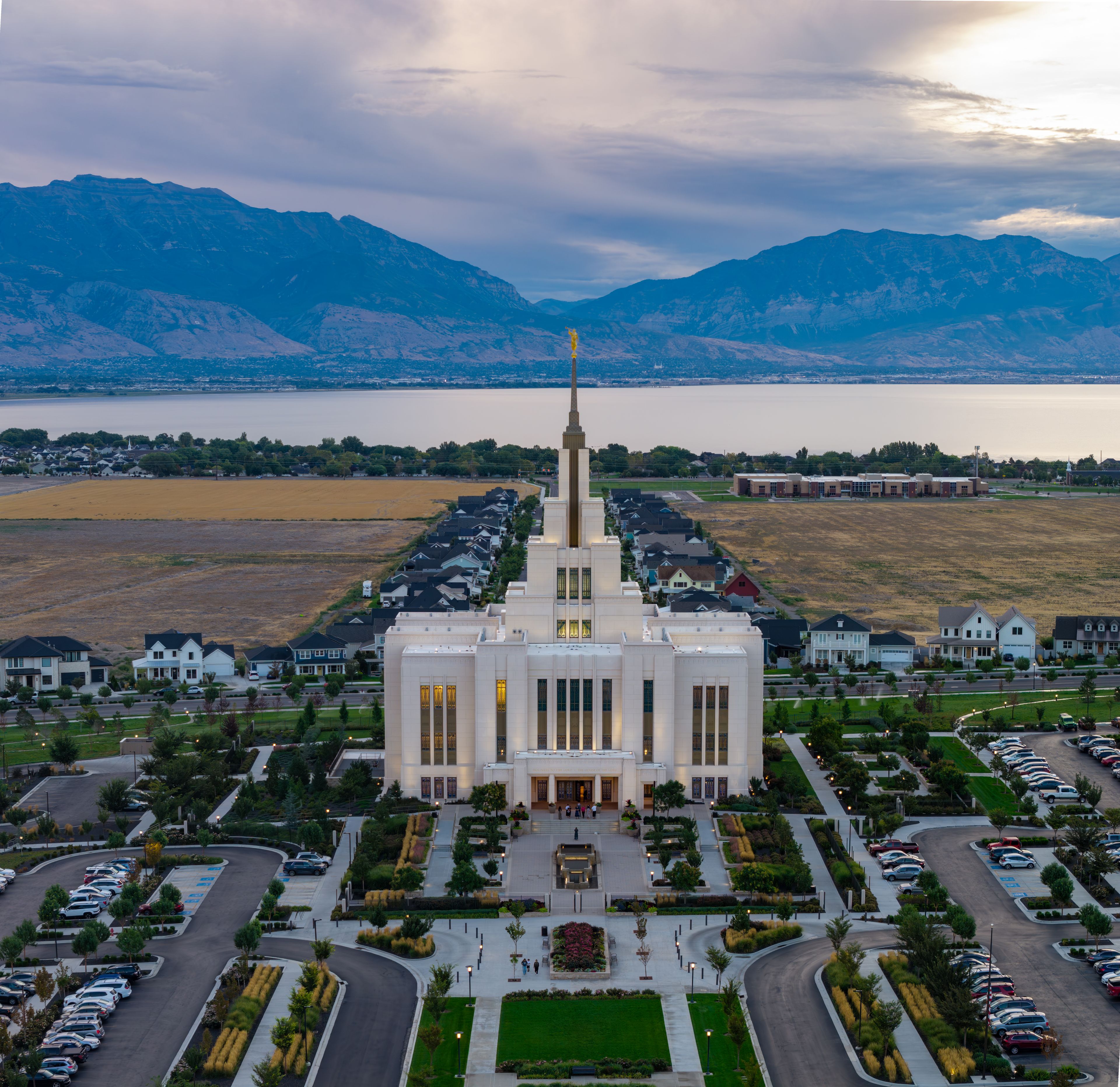 A large white building with a tall spire, surrounded by a parking lot and residential houses, with mountains and a lake in the background.