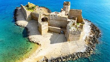 A coastal fortress with a tower, surrounded by clear blue waters and rocky terrain.