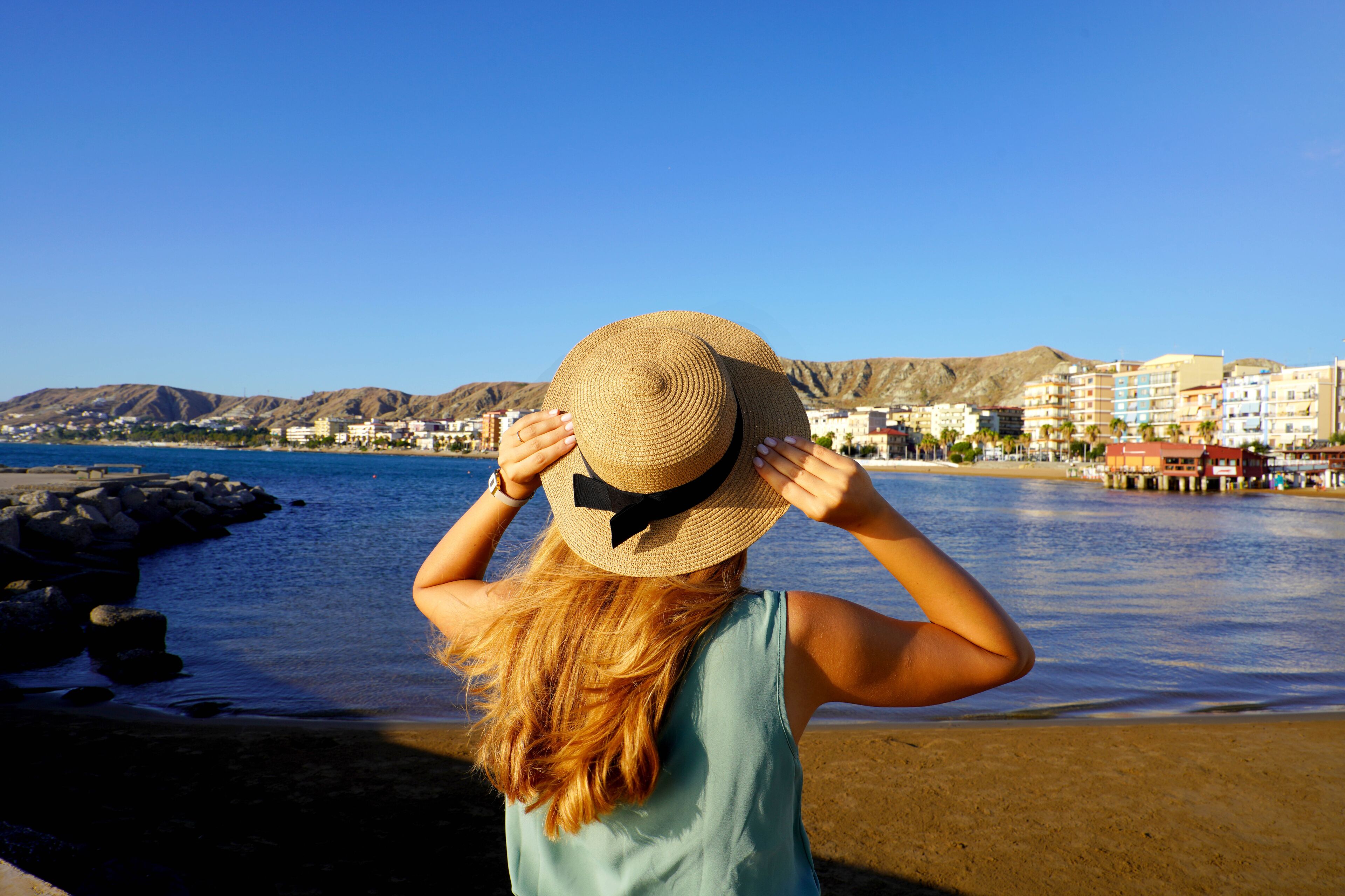 Sunset back view of girl holding straw hat in Crotone city on Calabria coast, Italy