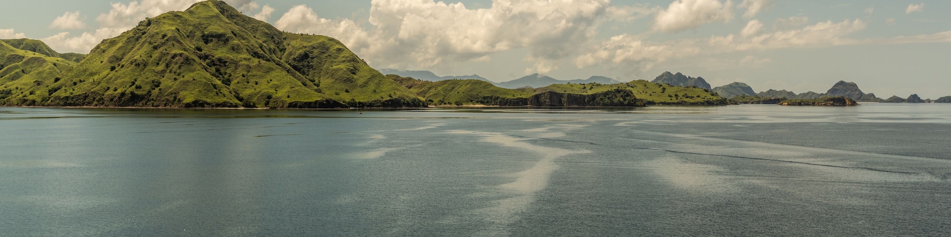 Rinca Island, Indonesia - February 24, 2019: Hills rolling down to sea on Westside coast in Savu Sea under cloudscape with white patches. Green hills in distance and flat sea.