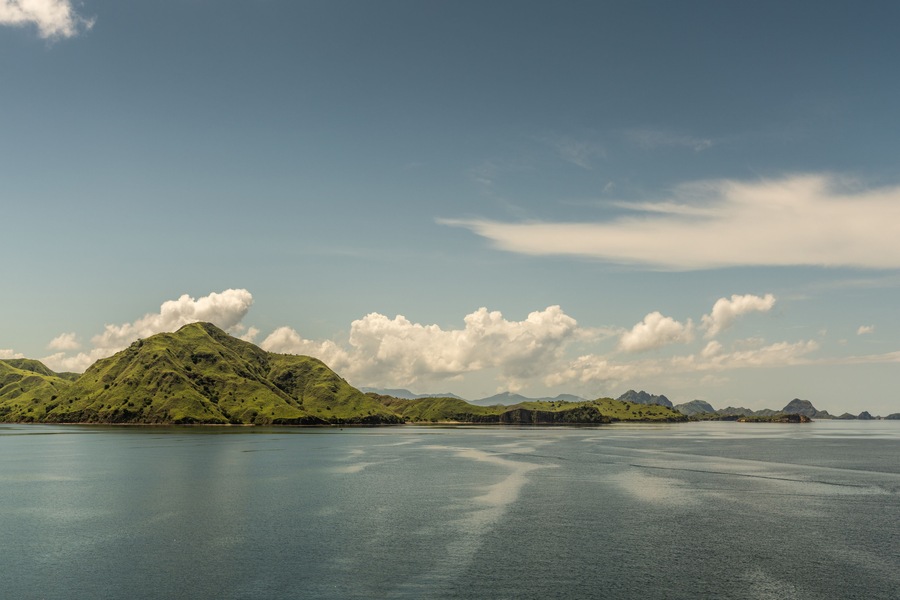 Rinca Island, Indonesia - February 24, 2019: Hills rolling down to sea on Westside coast in Savu Sea under cloudscape with white patches. Green hills in distance and flat sea.