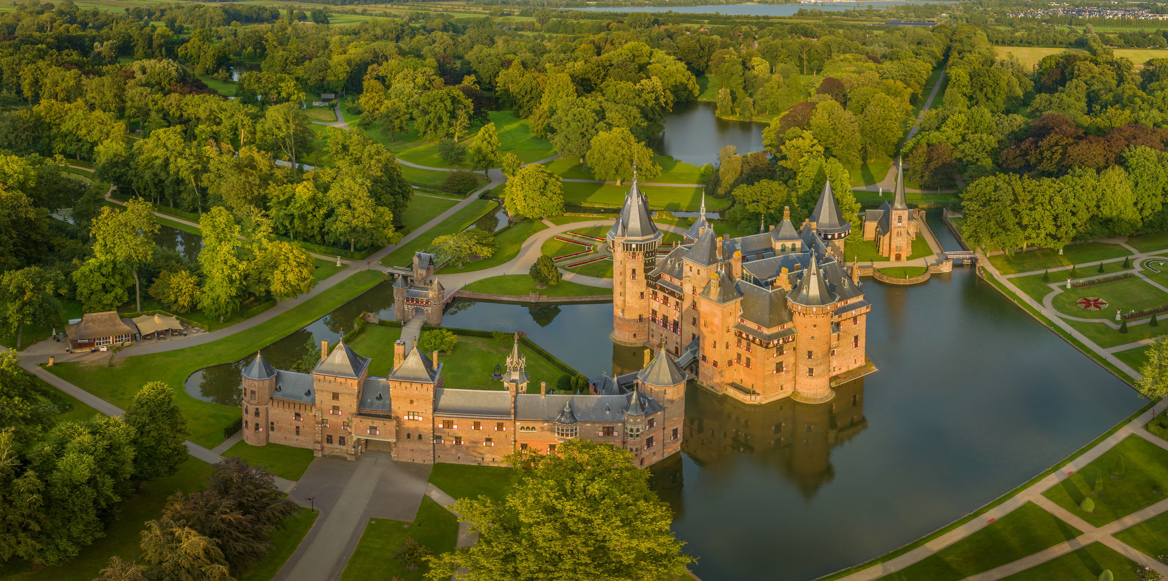 Un grand château entouré de douves, d’une végétation luxuriante et d’une rivière.