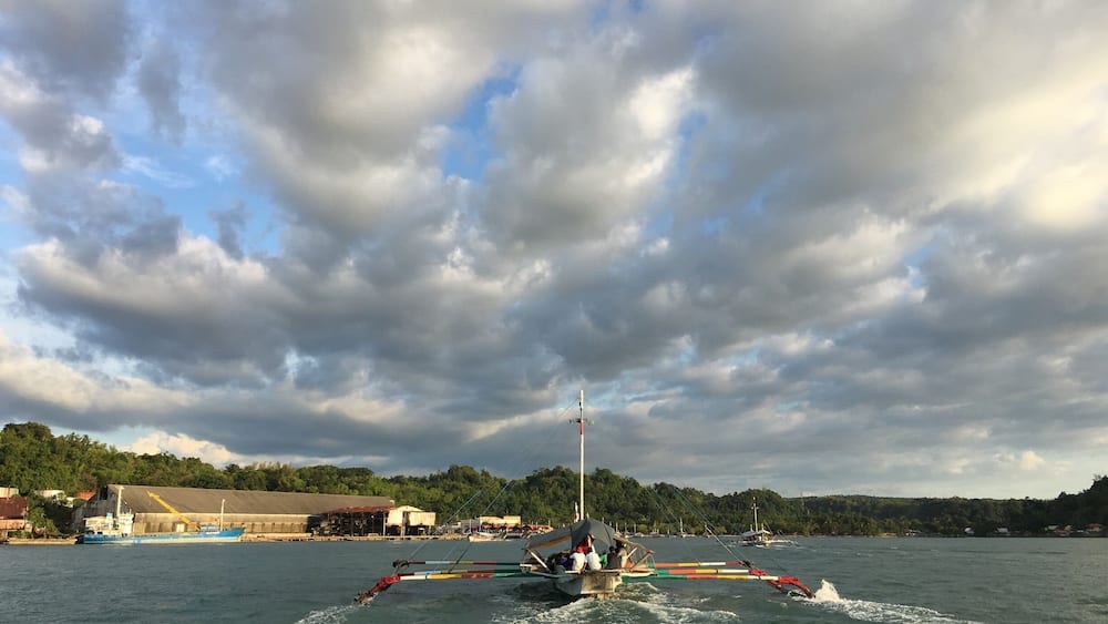 A traditional boat with colorful sails on a body of water, a docked ship in the background, and a cloudy sky.
