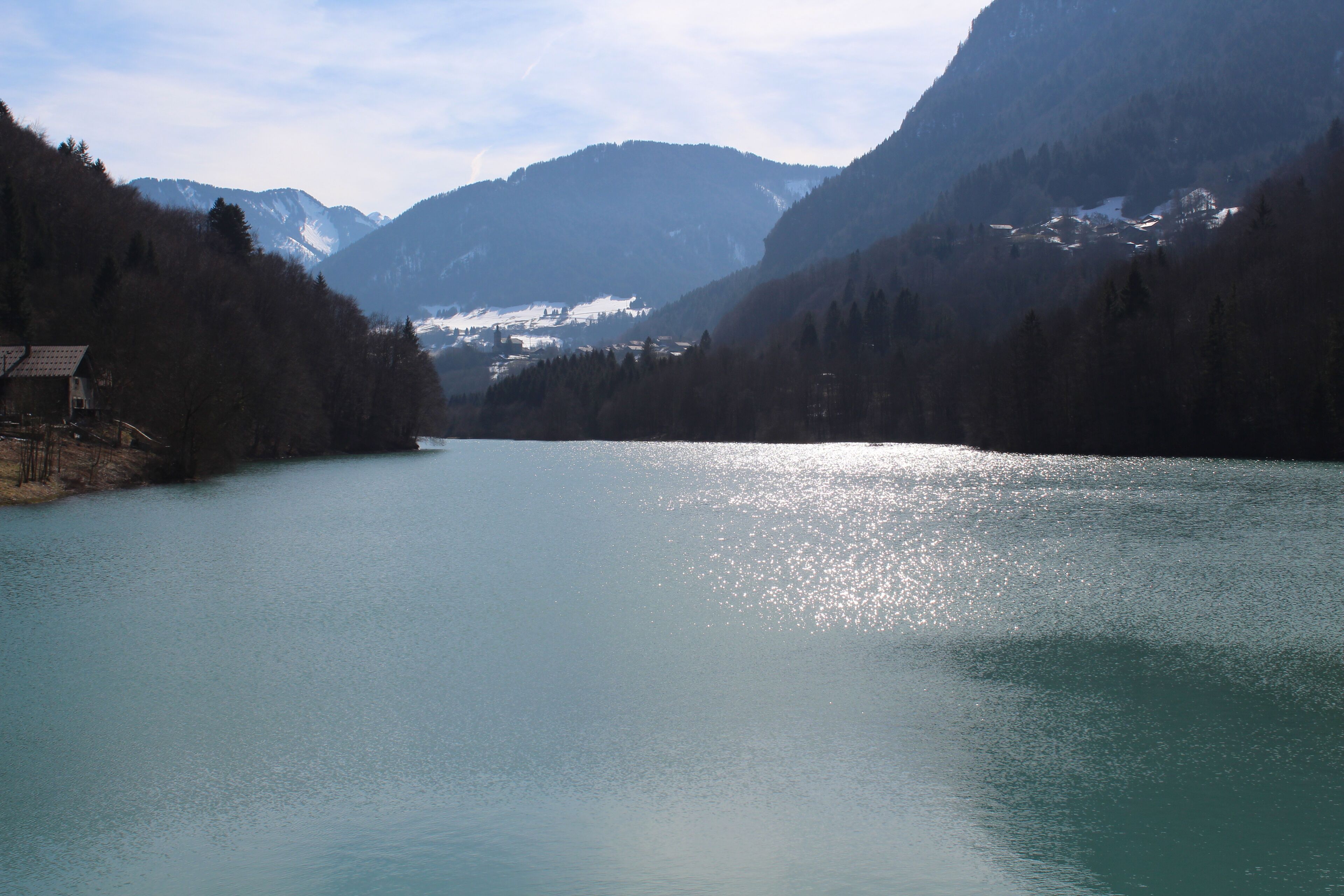 A calm lake surrounded by forested mountains with a small house on the left.