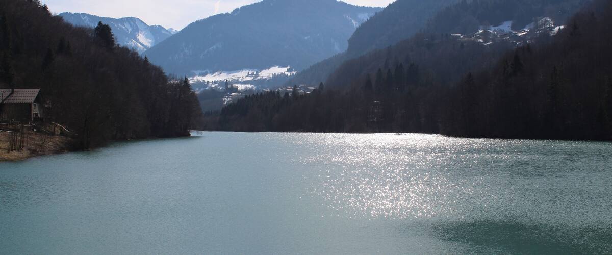 A calm lake surrounded by forested mountains with a small house on the left.