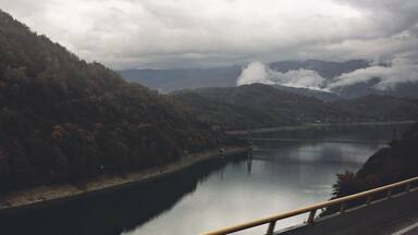A river with a bridge, surrounded by hills and cloudy skies.