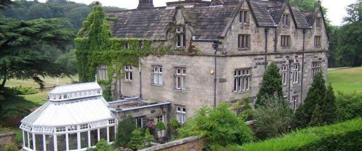 A large stone building with a glass conservatory, surrounded by lush greenery.