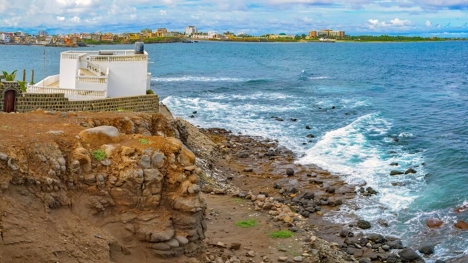 Una costa rocosa con olas rompiendo sobre la playa, un edificio blanco con un balcón con vistas al agua y un paisaje urbano distante con edificios y un cielo despejado.