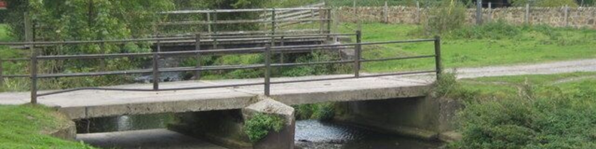 A concrete bridge over a narrow canal with a metal railing, surrounded by greenery and trees.
