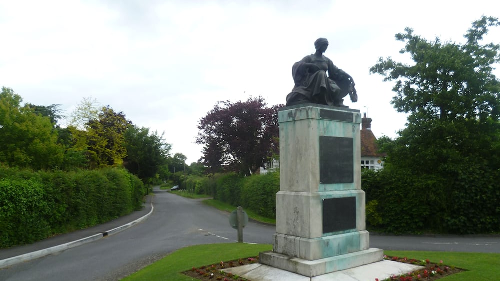 A statue on a pedestal in a grassy area beside a road.