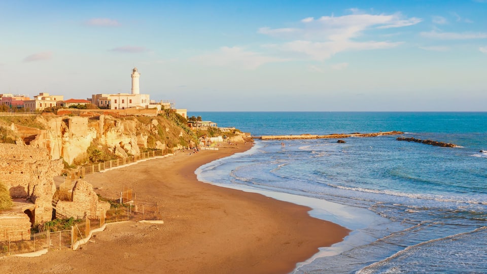 A coastal landscape with a lighthouse, sandy beach, and rocky cliffs.