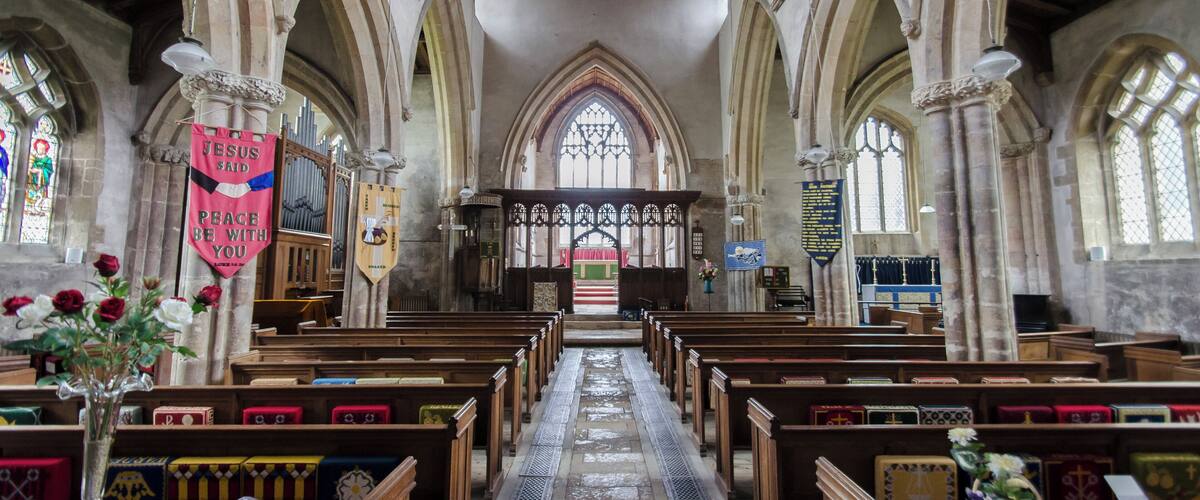 A church interior with wooden pews, a central aisle, stained glass windows, and religious banners.