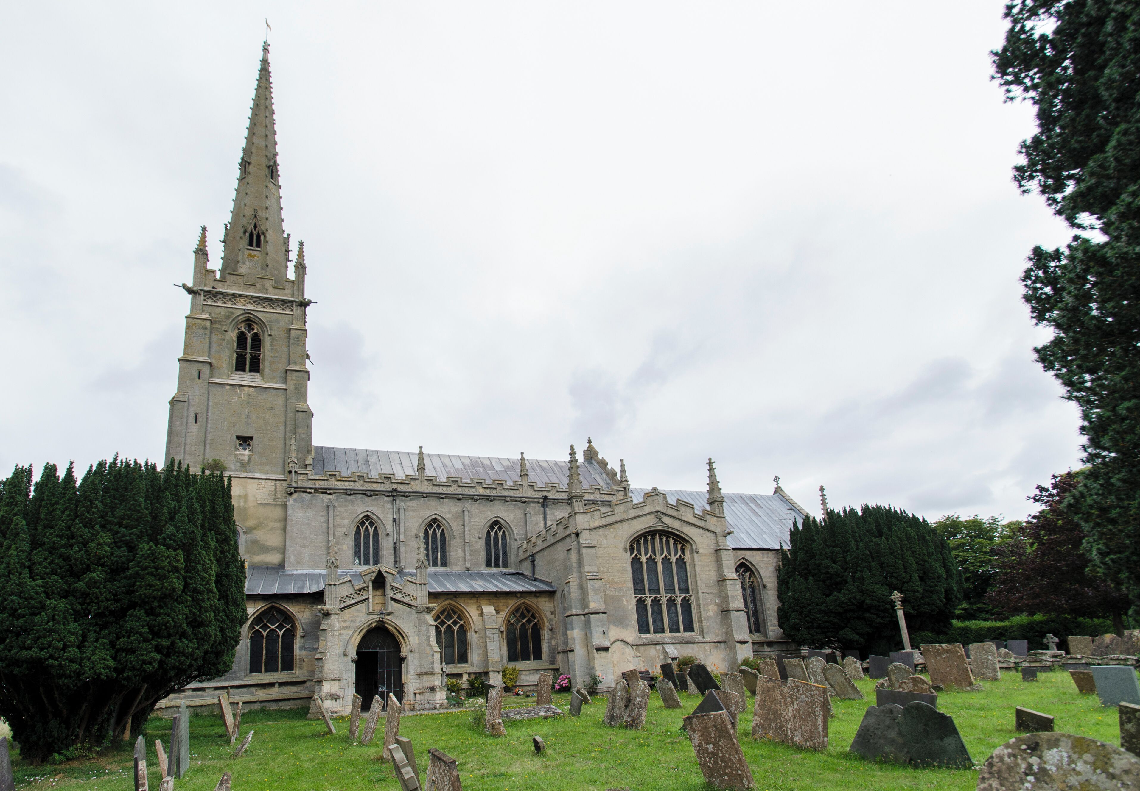 The church dates from circa 1300, and is built from ashlar with the lead roofs. The church is a cruciform plan with a west tower and spire, nave with north and south aisles, south porch, transepts, chancel and north vestry. The tower is of four stages, the two lower of circa 1300, the two upper of circa 1500. On the east side of the nave the original steeply pitched roofline is visible. The tower has ornate pinnacles and projecting gargoyles, the octagonal spire is 15th century with lucarnes, Crockets and finial. The nave has clerestory windows of three lights. There are moulded eaves with corbel heads above,and a frieze of shields, battlements and ornate pinnacles. The north aisle has an early 14th century west window and on the roof is a small bell turret,the north transept has early 14th century window of three lights. The chancel is 15th century and was restored in the 19th C. It has two large windows on both north and south sides each of three lights. There are hood moulds and grotesque animal label stops.There is a large pointed east window of five lights with perpendicular tracery. The original roofline of the chancel is visible on the east end of the nave. The south transept was probably remodelled in the 15th century from a detached lady Chapel built circa 1275. The west side of the transept has 14th century windows with head label stops. The porch is 14th century and has 15th century ornamentation, it has projecting gargoyles and ornate pinnacles. It contains stone benches and a corbel that once supported a roof, one is dated 1631. The 14th century west door has foliated capitals with elaborate finials. The nave has three bays, with piers from circa 1300. The south wall of the south transept has three 13th century sedilia and two aumbries. The chancel arch is from the 14th century and has caryatid figures. There is a 14th century screen which was restored in the 19th century. There is a 14th century hexagonal font with ornate Gables and pinnacles. The church contains various small 18th and 19th-century monuments. The church has recently undergone much restoration work including work to the spire, tower, bells, roof, windows and a new boiler system.
