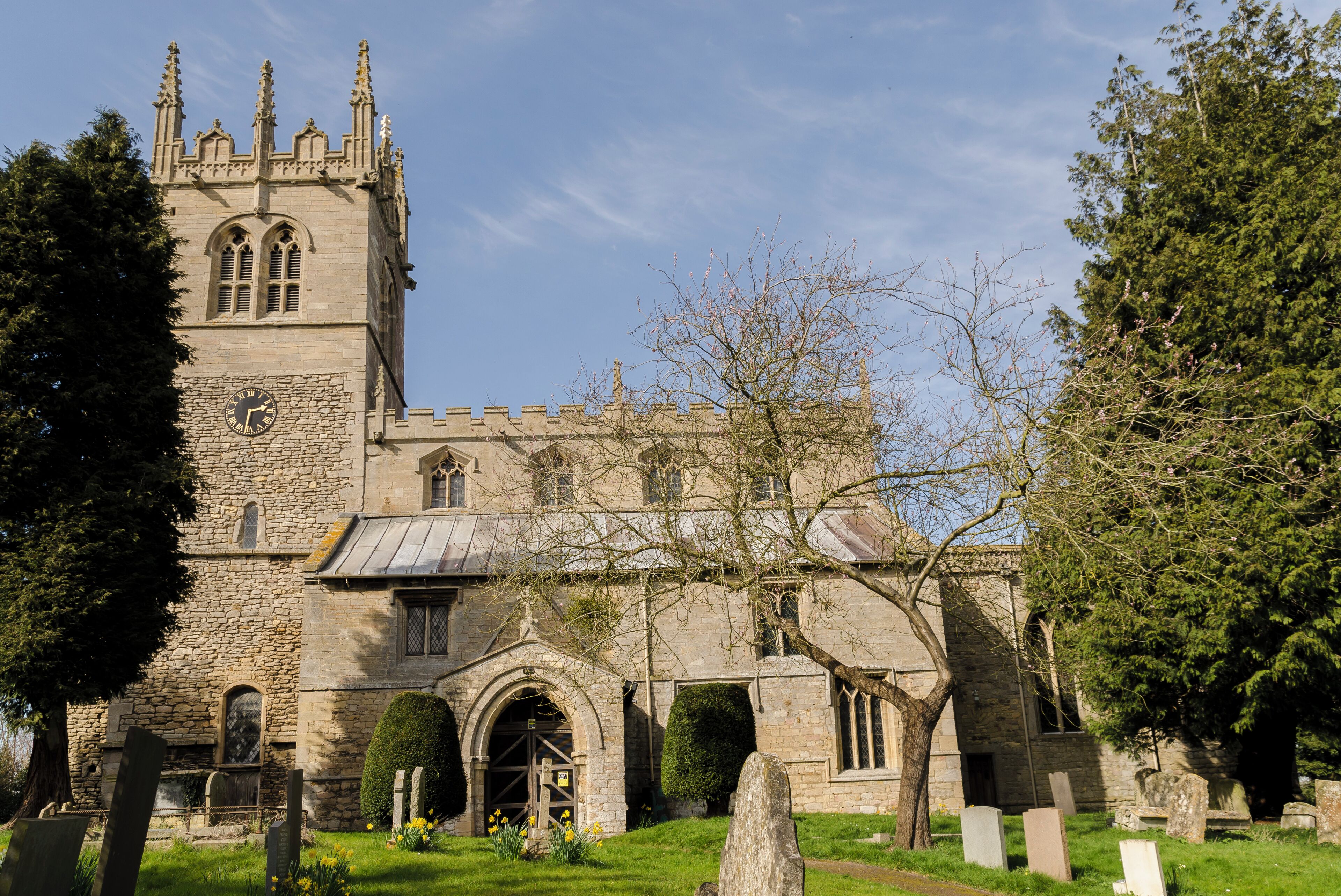 A historic stone church with a clock tower, surrounded by a cemetery with headstones and flowering plants.