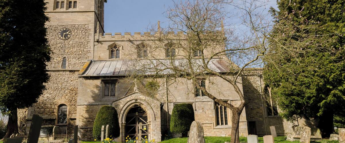 A historic stone church with a clock tower, surrounded by a cemetery with headstones and flowering plants.