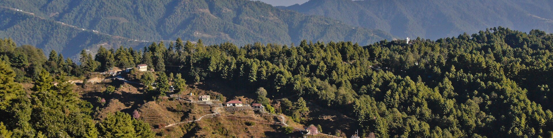 Paisagem montanhosa com picos nevados ao fundo e um vale com casas dispersas e florestas densas em primeiro plano.