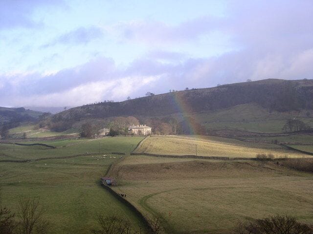 A rural landscape with rolling hills, a rainbow, and a building in the distance.