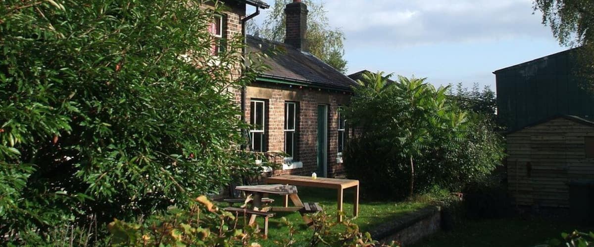 A brick house with a green roof, surrounded by lush greenery and a wooden shed.