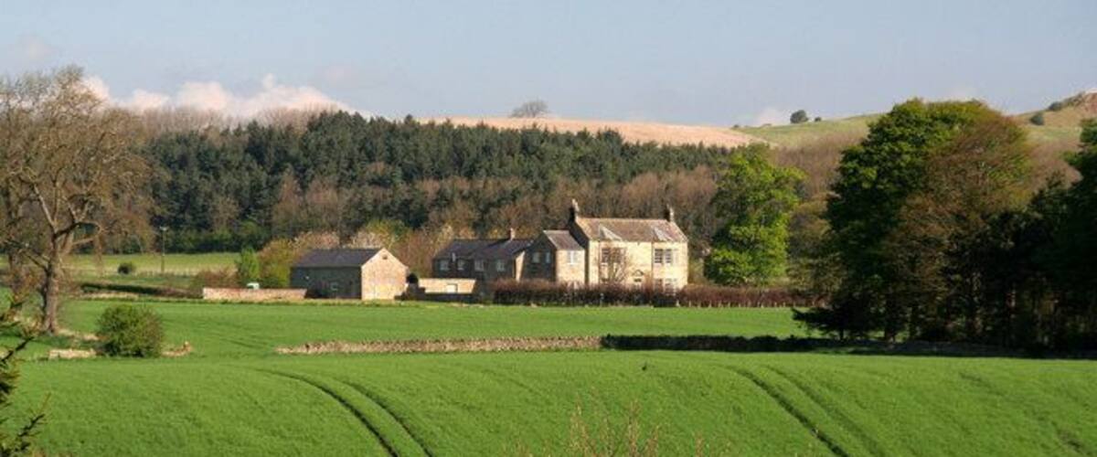 A rural landscape with rolling green fields, a stone house, and a forested hill in the background.