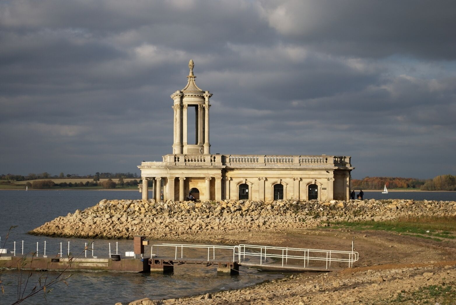 A classical stone structure with a central tower, situated on a rocky shoreline by a body of water.