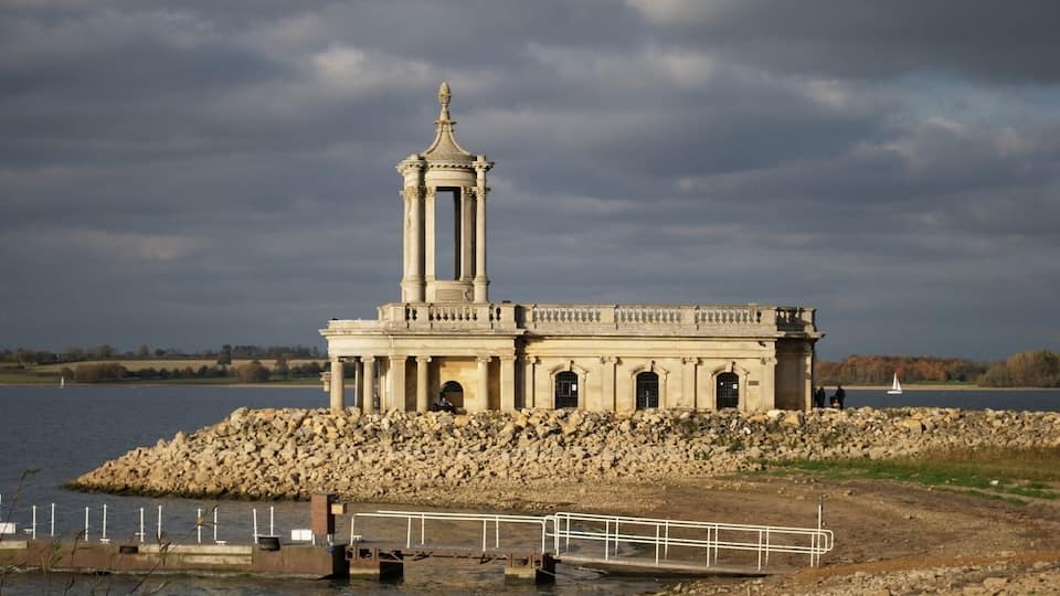 A classical stone structure with a central tower, situated on a rocky shoreline by a body of water.